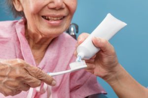 Older woman holding a toothbrush as someone puts toothpaste on it